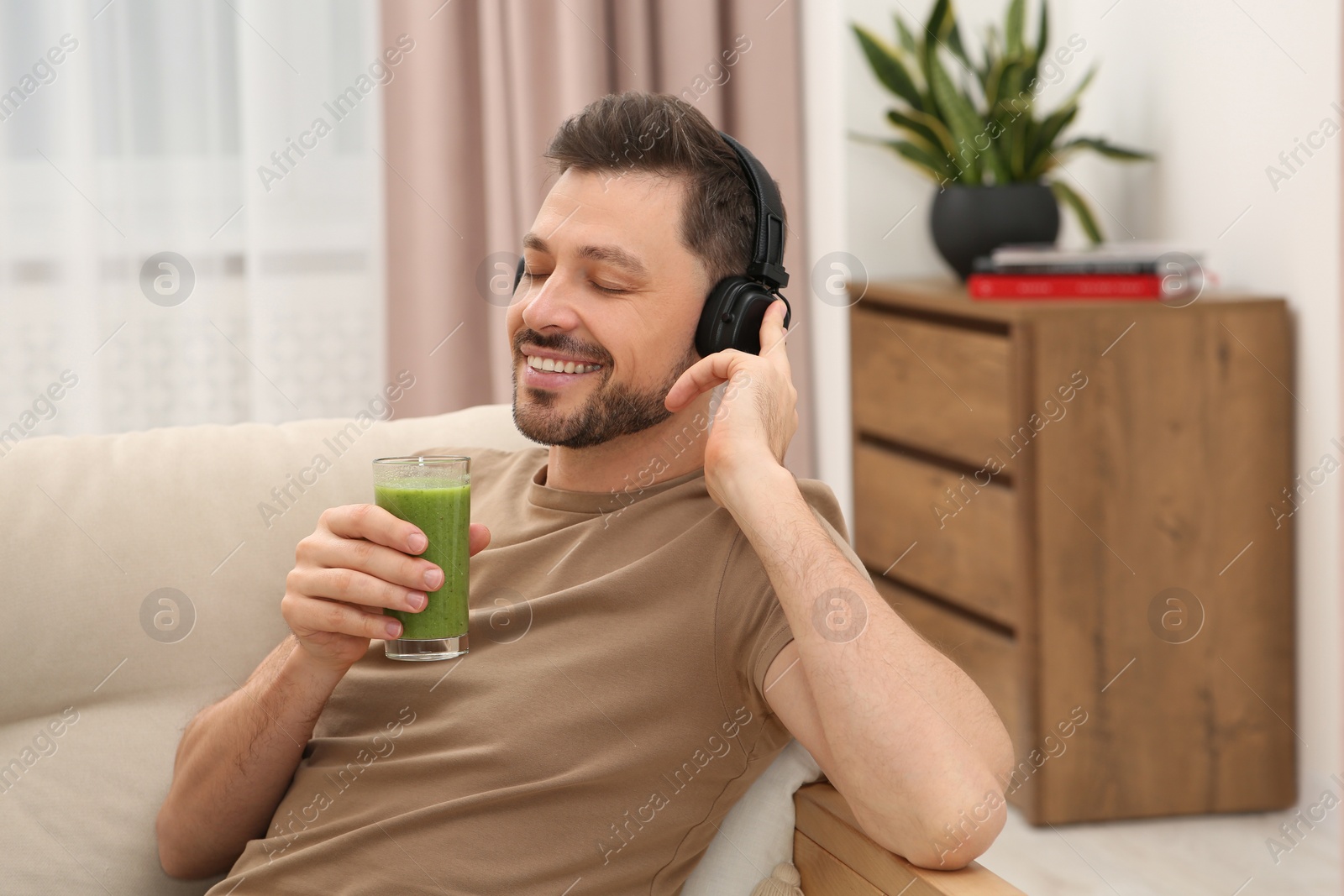 Photo of Happy man in headphones holding glass of delicious smoothie and listening to music at home