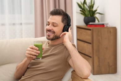 Photo of Happy man in headphones holding glass of delicious smoothie and listening to music at home