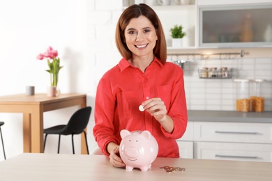 Photo of Woman putting coin into piggy bank at table indoors. Saving money