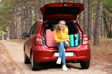 Photo of Beautiful young woman sitting in car trunk loaded with suitcases on forest road