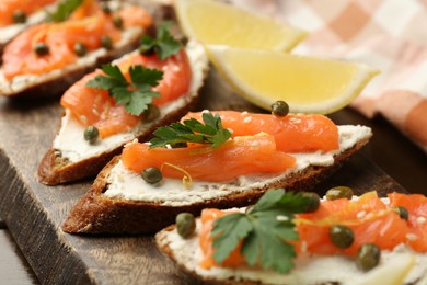 Photo of Tasty canapes with salmon, capers, lemon and cream cheese on wooden board, closeup