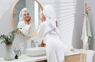Photo of Beautiful woman in white robe applying cream near mirror in bathroom