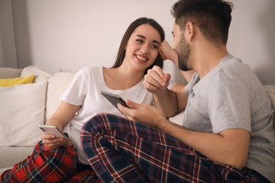 Happy couple in pajamas with gadgets on sofa at home