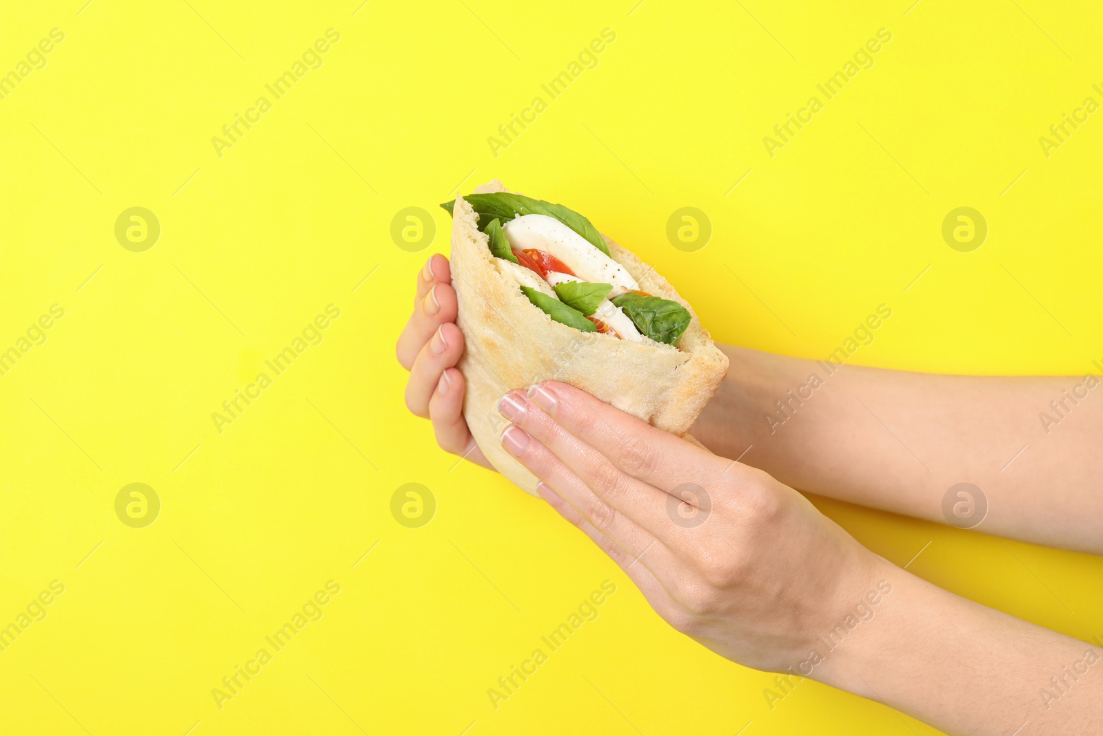 Photo of Woman holding delicious pita sandwich with mozzarella, tomatoes and basil on yellow background, closeup