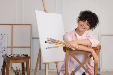 Young woman holding brushes near easel with canvas in studio