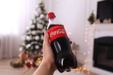 Photo of MYKOLAIV, UKRAINE - JANUARY 13, 2021: Woman holding bottle of Coca-Cola in room with Christmas tree, closeup