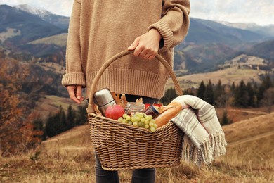 Image of Woman holding wicker picnic basket with thermos, snacks and plaid in mountains on autumn day, closeup
