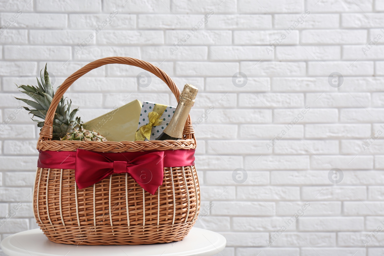 Photo of Wicker gift basket with pineapple and bottle of champagne on table near white brick wall. Space for text