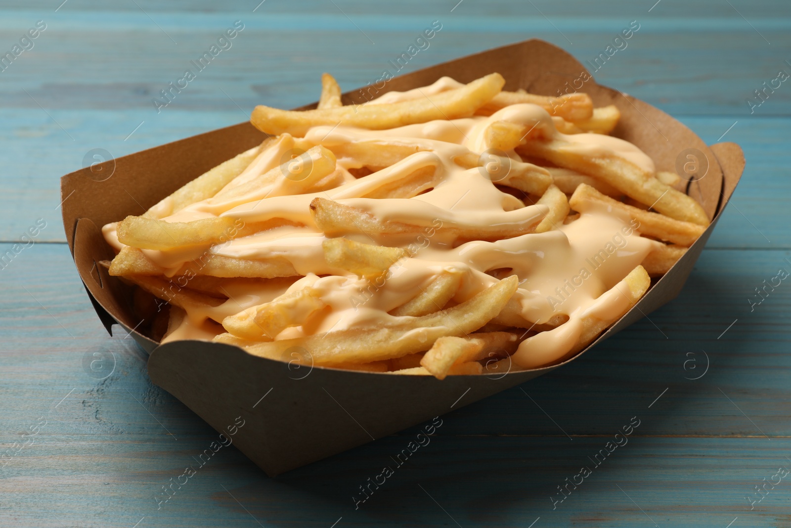 Photo of Tasty potato fries and cheese sauce in paper container on light blue wooden table, closeup