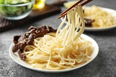 Chopsticks with tasty cooked rice noodles over plate on grey table