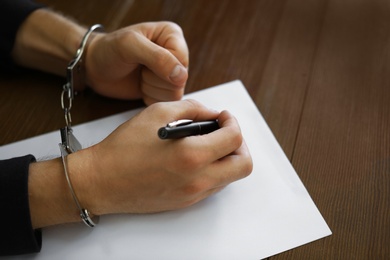 Photo of Criminal in handcuffs writing confession at desk, closeup