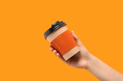 Woman holding takeaway paper coffee cup with cardboard sleeve on orange background, closeup