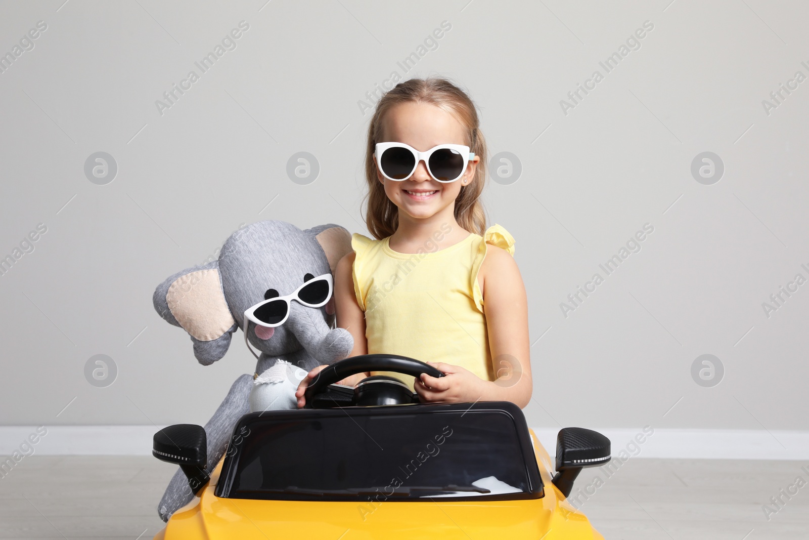 Photo of Cute little girl with toy elephant driving children's car near grey wall indoors