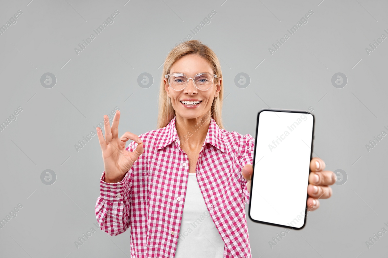 Photo of Happy woman holding smartphone with blank screen and showing OK gesture on grey background