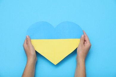 Photo of Woman holding paper heart in colors of Ukrainian flag on light blue background, top view