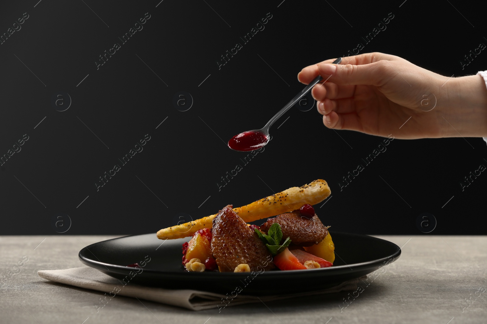 Photo of Food stylist adding sauce to delicious dish with chicken, parsnip and strawberries for photoshoot at grey table in studio, closeup