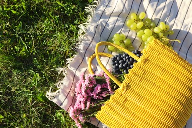 Yellow wicker bag with beautiful flowers, grapes and blueberries on picnic blanket outdoors, top view