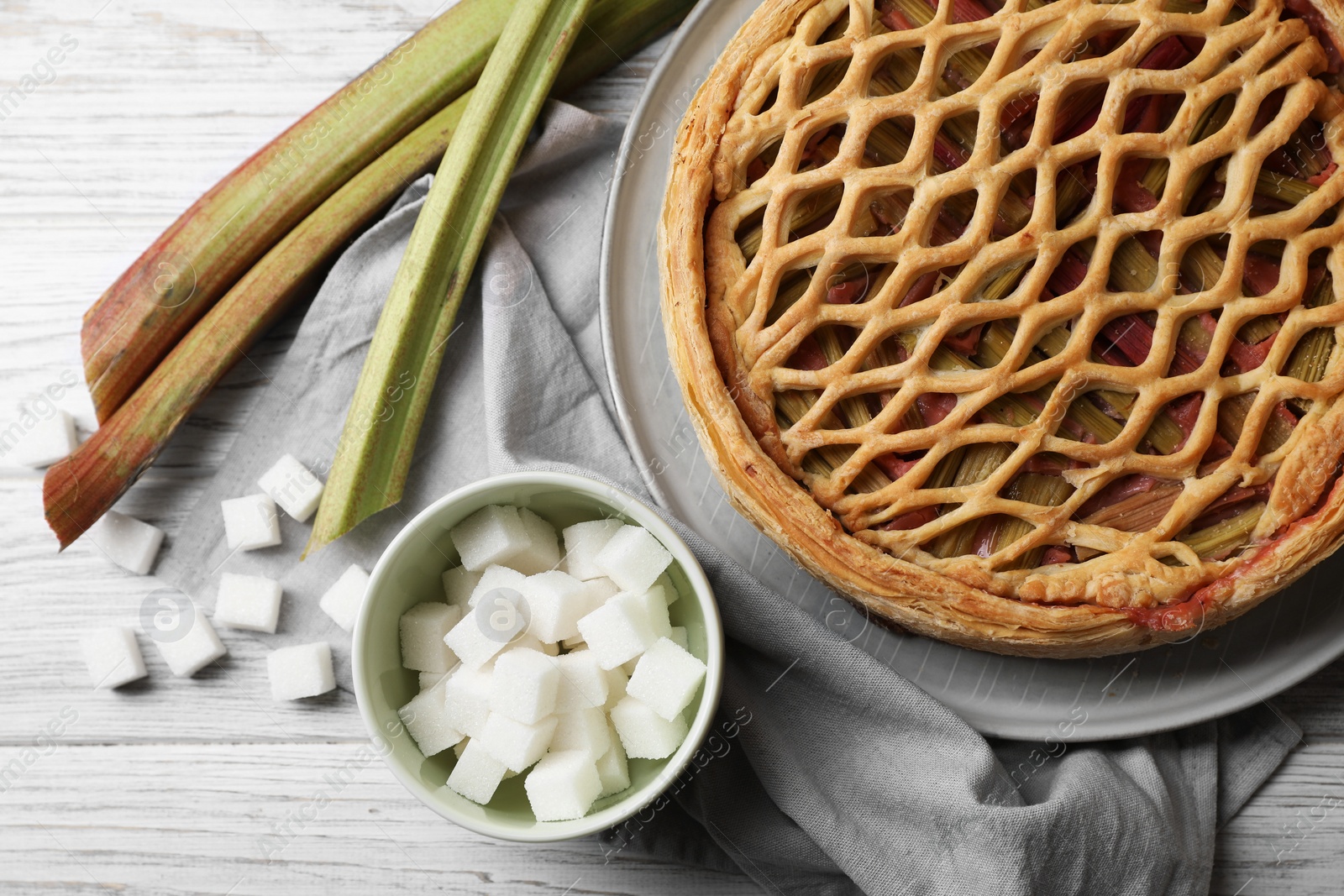 Photo of Freshly baked rhubarb pie, stalks and sugar cubes on light wooden table, flat lay