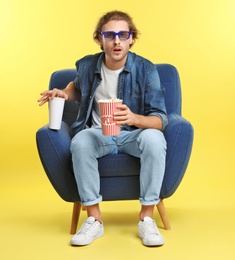 Photo of Emotional man with 3D glasses, popcorn and beverage sitting in armchair during cinema show on color background