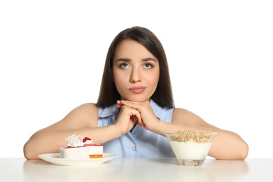 Doubtful woman choosing between yogurt with granola and cake at table on white background