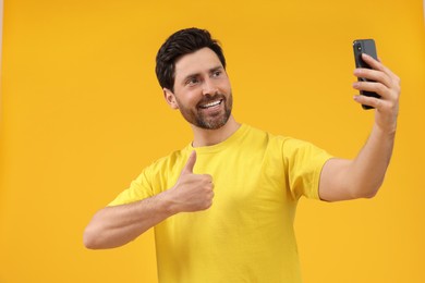 Photo of Smiling man taking selfie with smartphone and showing thumbs up on yellow background