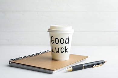 Photo of Paper coffee cup with phrase GOOD LUCK and stationery on white wooden table