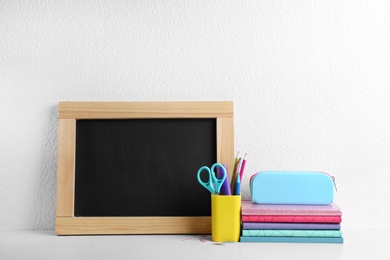 Photo of Blank small chalkboard and different school stationery on wooden table near white wall. Space for text