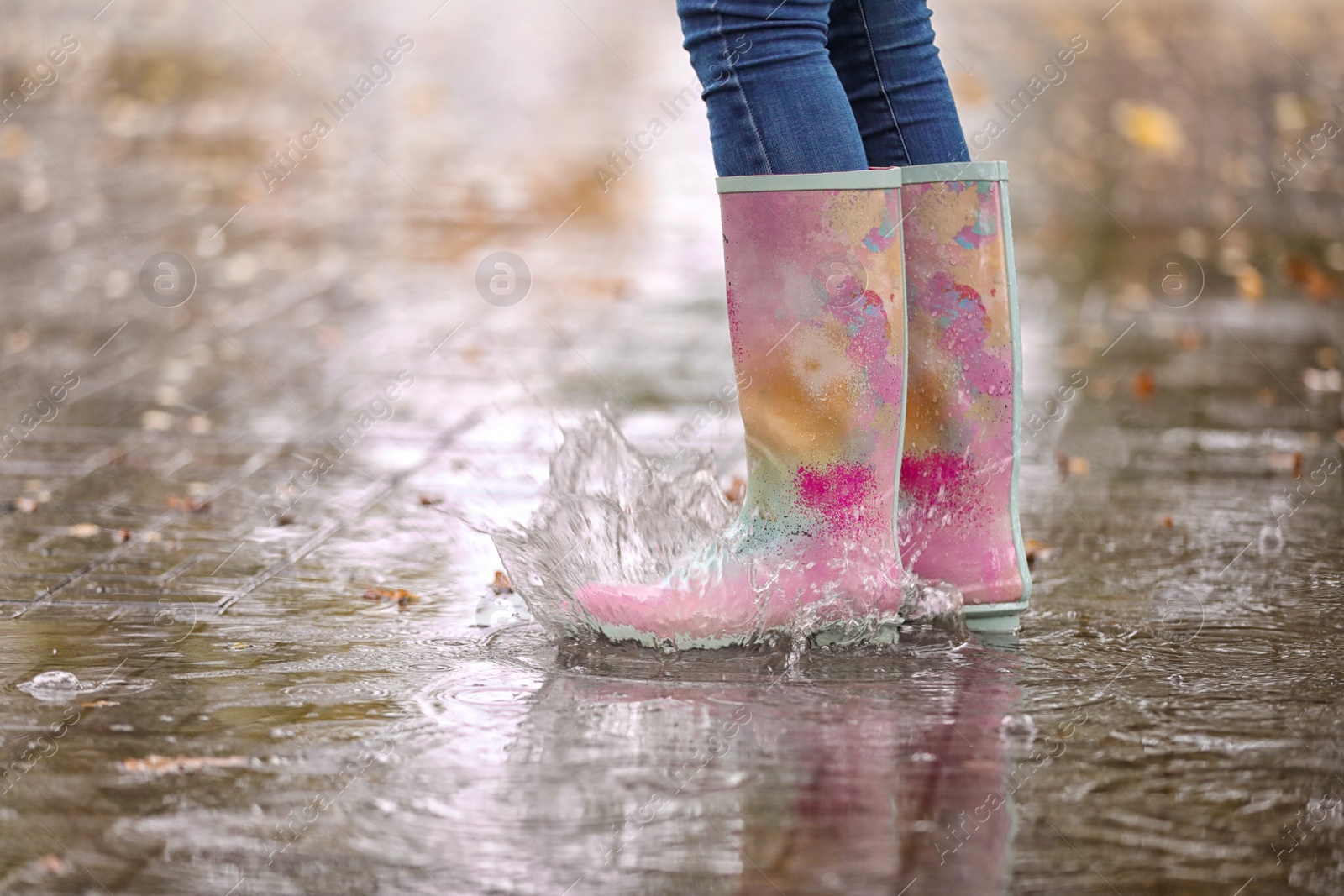 Photo of Woman wearing rubber boots splashing in puddle after rain, focus on legs. Autumn walk