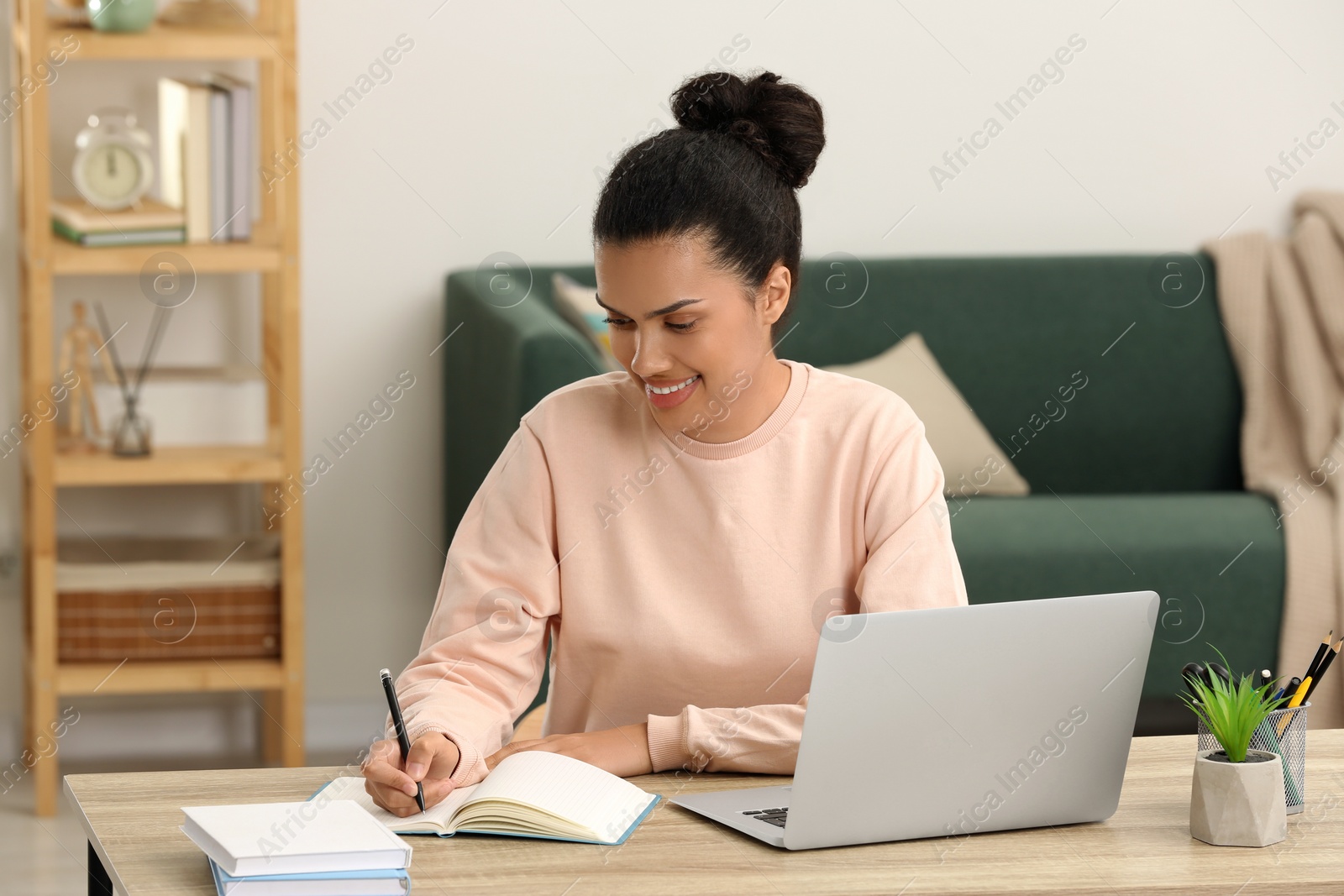 Photo of Beautiful African American woman writing in notepad near laptop at table in room