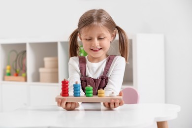 Photo of Cute little girl playing with stacking and counting game at white table indoors. Child's toy