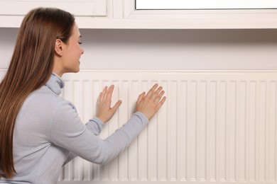 Woman warming hands on heating radiator indoors, space for text