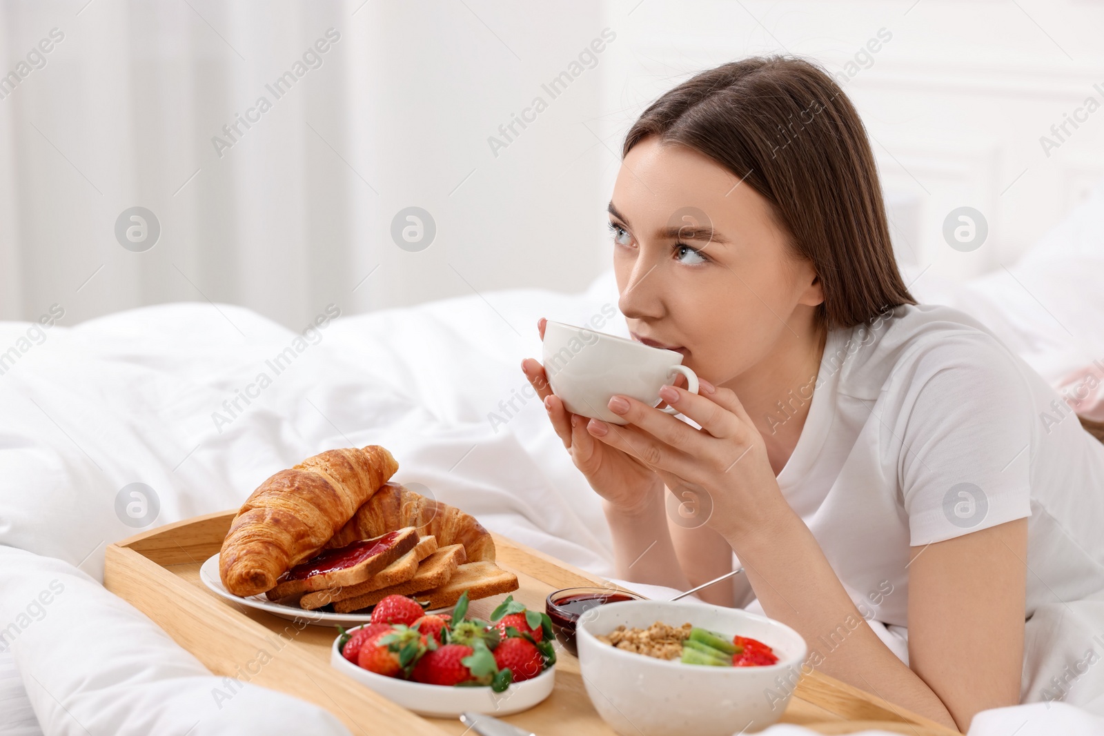 Photo of Beautiful woman drinking coffee near tray with breakfast on bed. Space for text