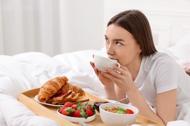 Beautiful woman drinking coffee near tray with breakfast on bed. Space for text