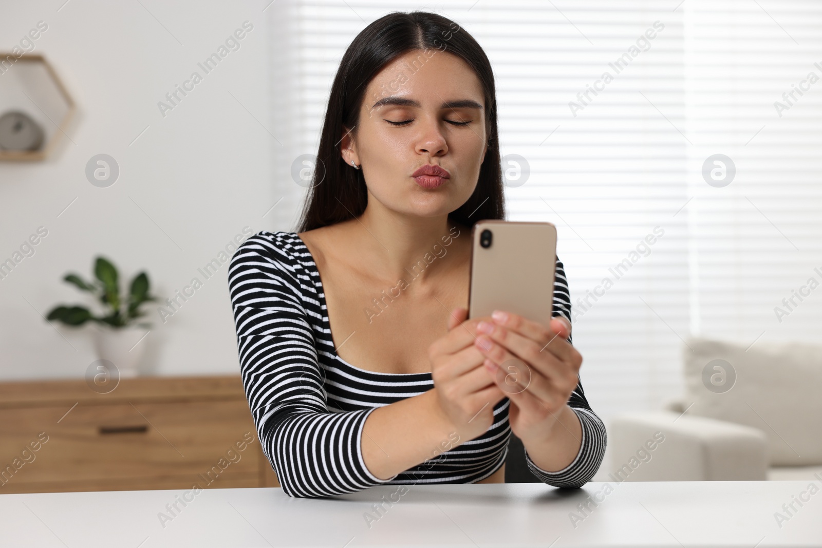 Photo of Young woman having video chat via smartphone and blowing kiss at table in room