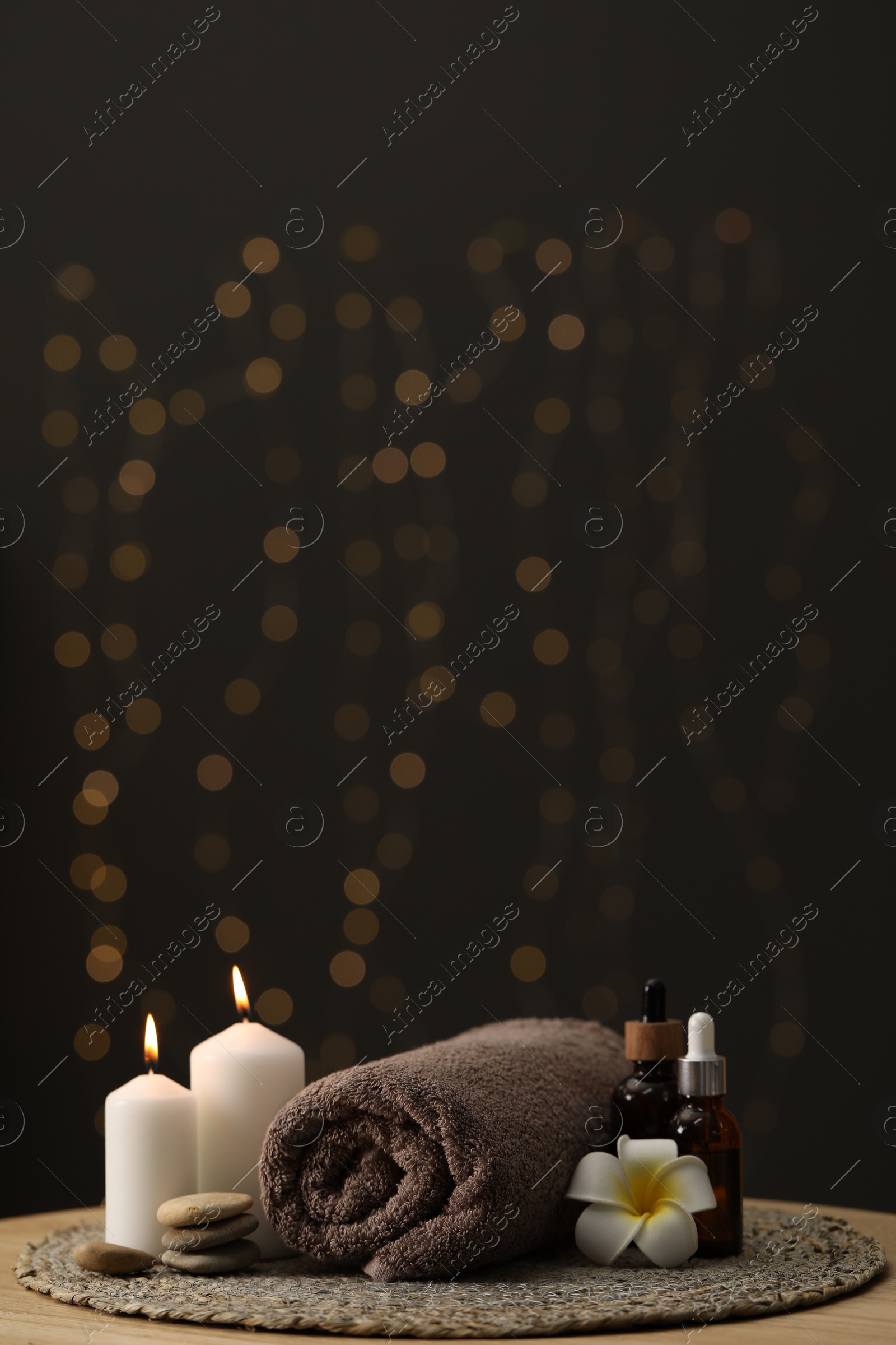 Photo of Spa composition. Rolled towel, cosmetic products, stones, burning candles and plumeria flower on table indoors. Space for text