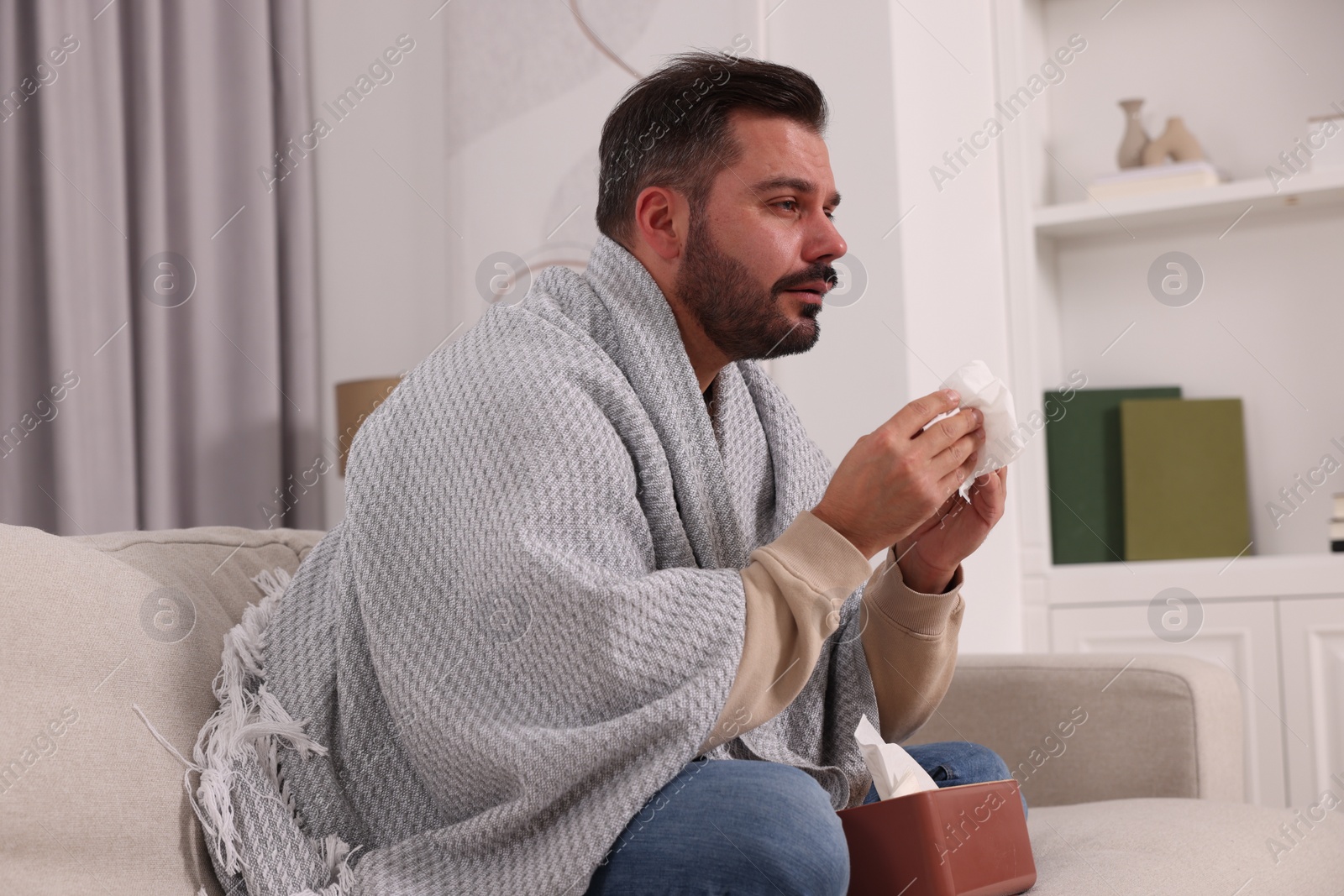 Photo of Sick man wrapped in blanket with tissues on sofa at home. Cold symptoms