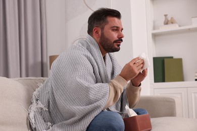 Sick man wrapped in blanket with tissues on sofa at home. Cold symptoms