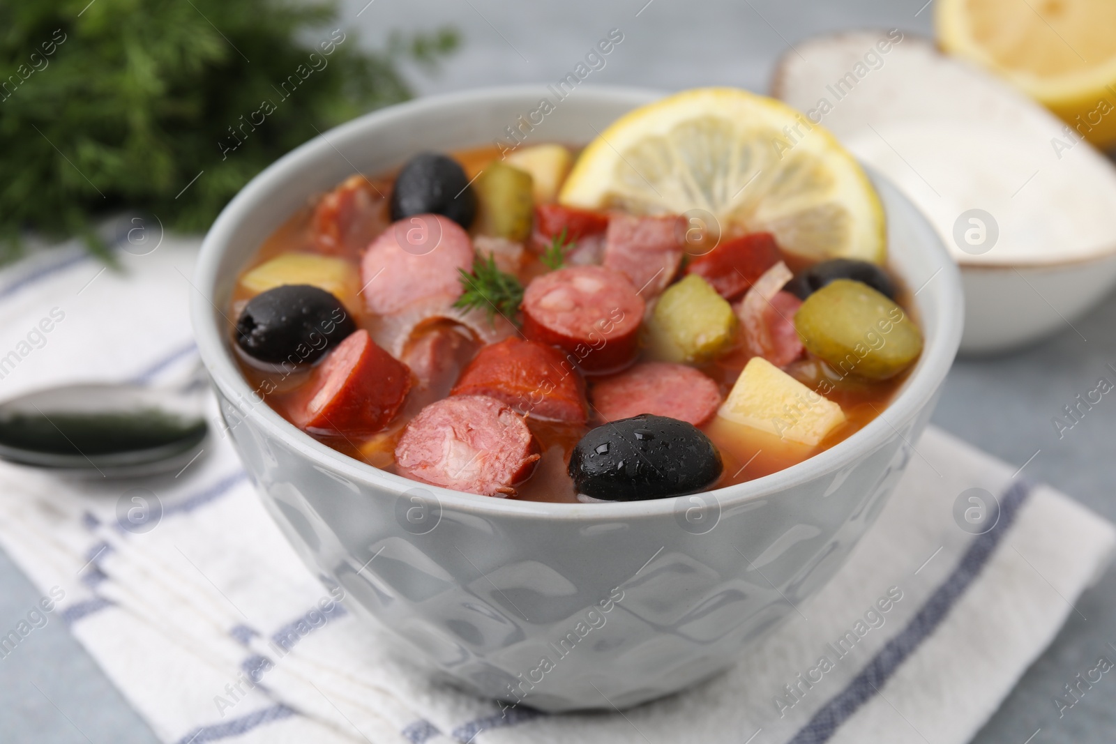 Photo of Meat solyanka soup with thin dry smoked sausages in bowl on grey table, closeup