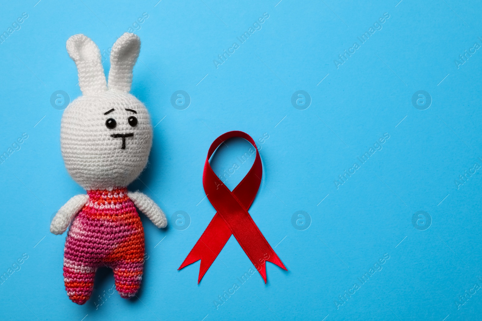 Photo of Cute knitted toy bunny and red ribbon on blue background, flat lay with space for text. AIDS disease awareness