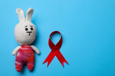 Photo of Cute knitted toy bunny and red ribbon on blue background, flat lay with space for text. AIDS disease awareness