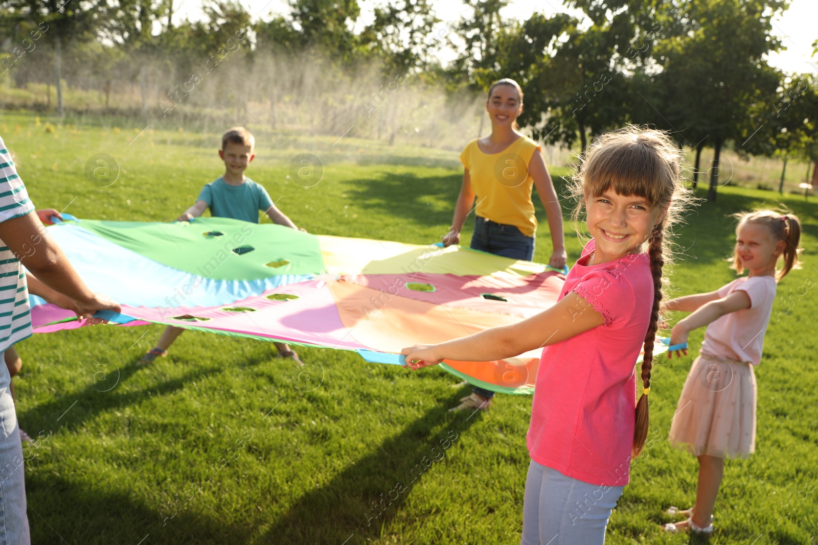 Photo of Group of children and teacher playing with rainbow playground parachute on green grass. Summer camp activity