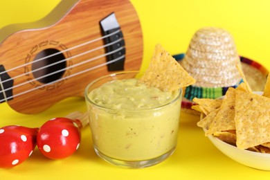 Delicious guacamole with nachos chips, Mexican sombrero hat, ukulele and maracas on yellow background, closeup