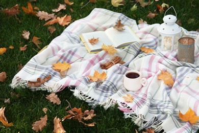 Plaid, cup of coffee, book, lantern and candle on green grass covered with autumn leaves