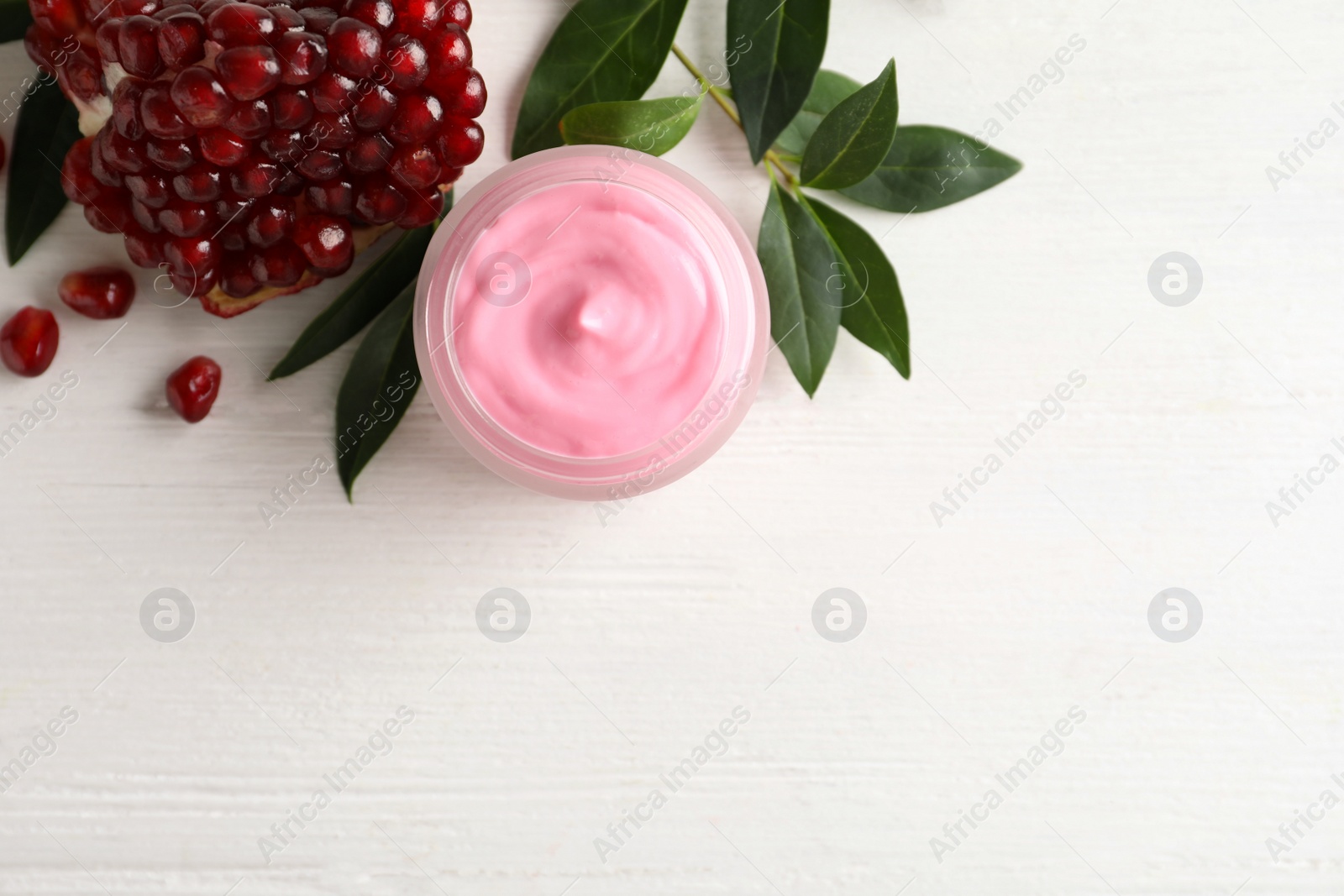 Photo of Jar with natural facial mask, pomegranate seeds and green leaves on white wooden table, flat lay. Space for text