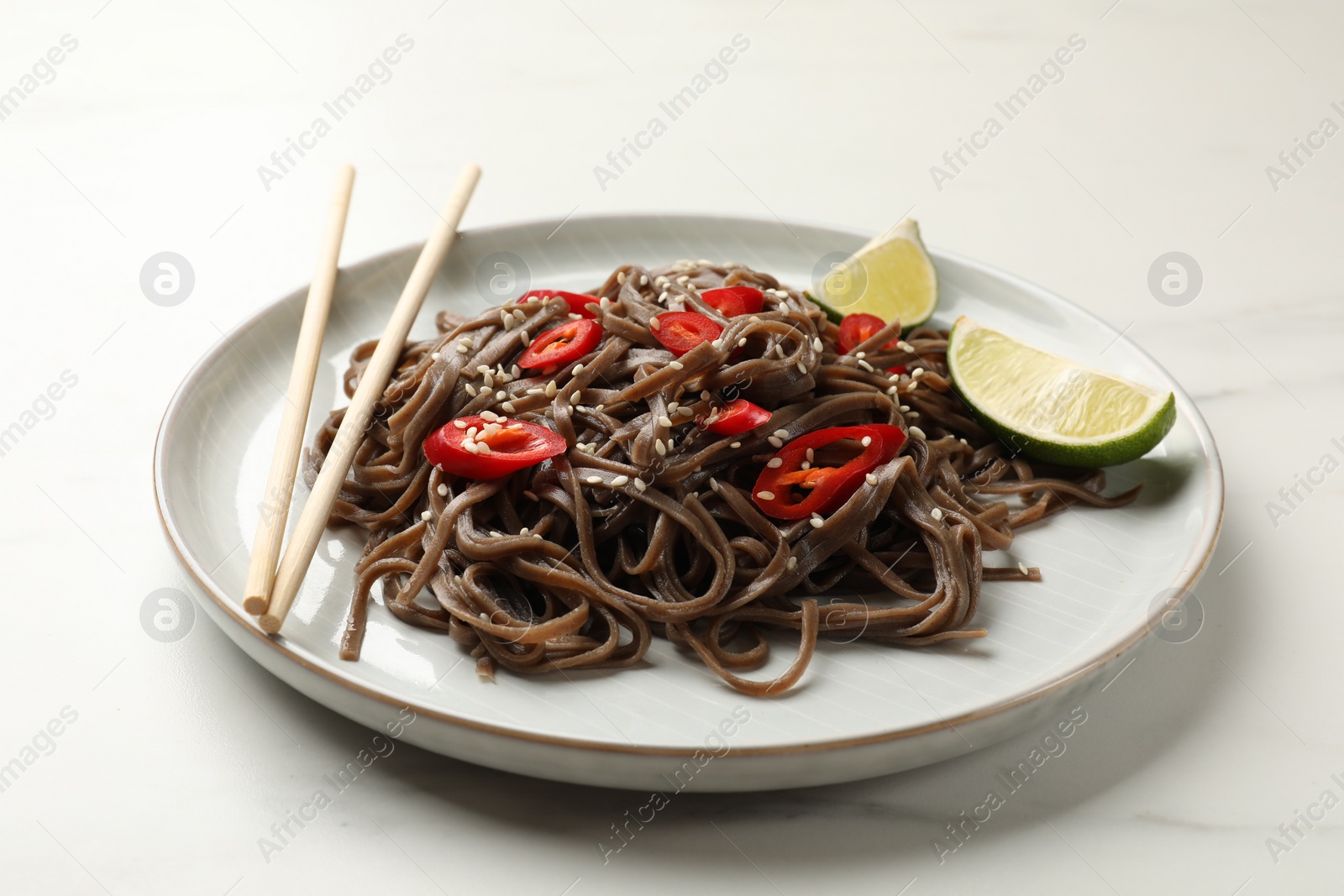 Photo of Tasty buckwheat noodles (soba) with chili pepper, lime and chopsticks on white marble table