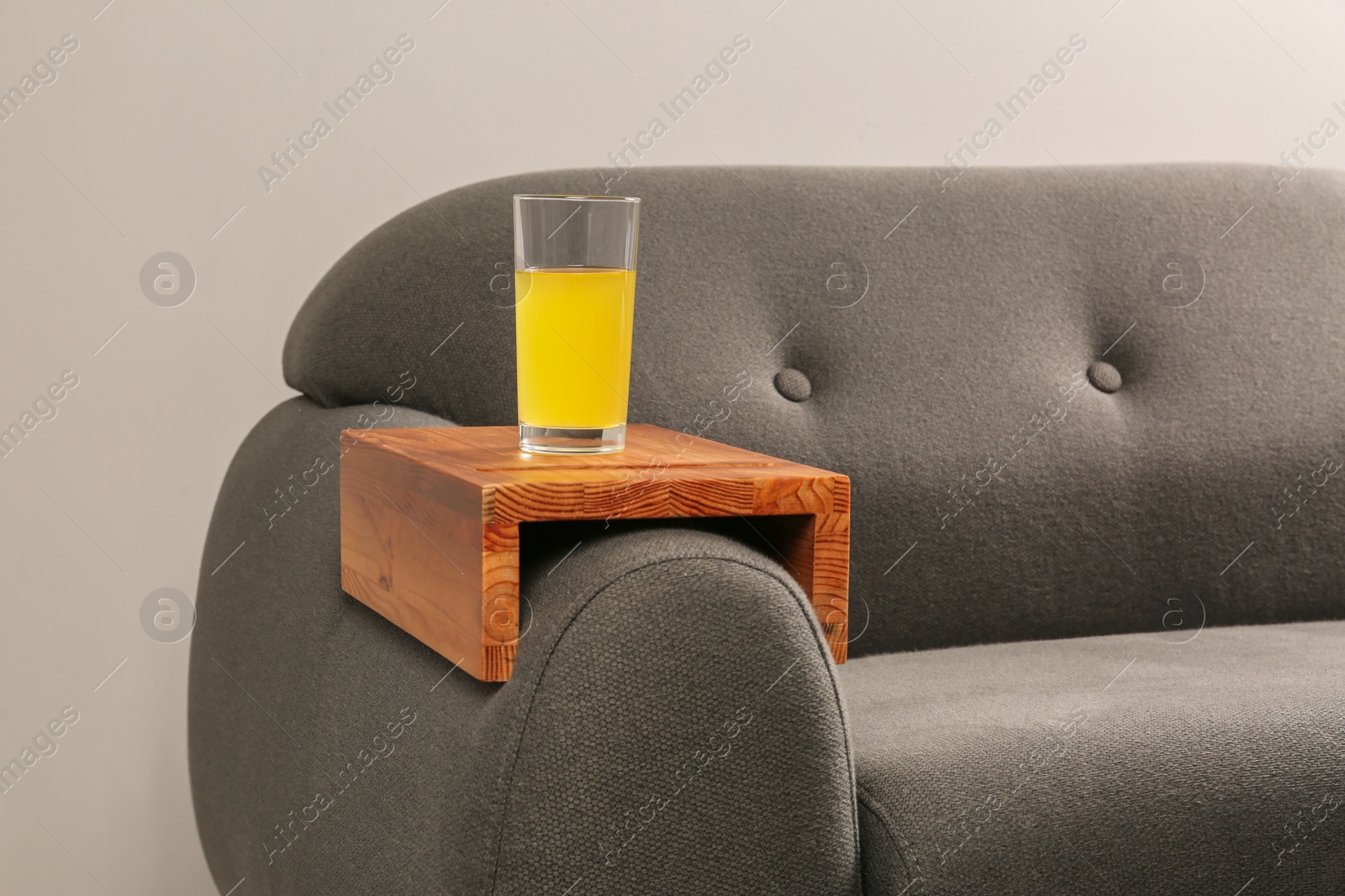 Photo of Glass of juice on sofa with wooden armrest table in room. Interior element