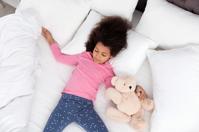 Photo of Cute little African-American girl with teddy bear sleeping in bed, top view