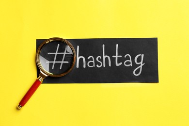 Photo of Black paper with word Hashtag, symbol and magnifying glass on yellow background, flat lay