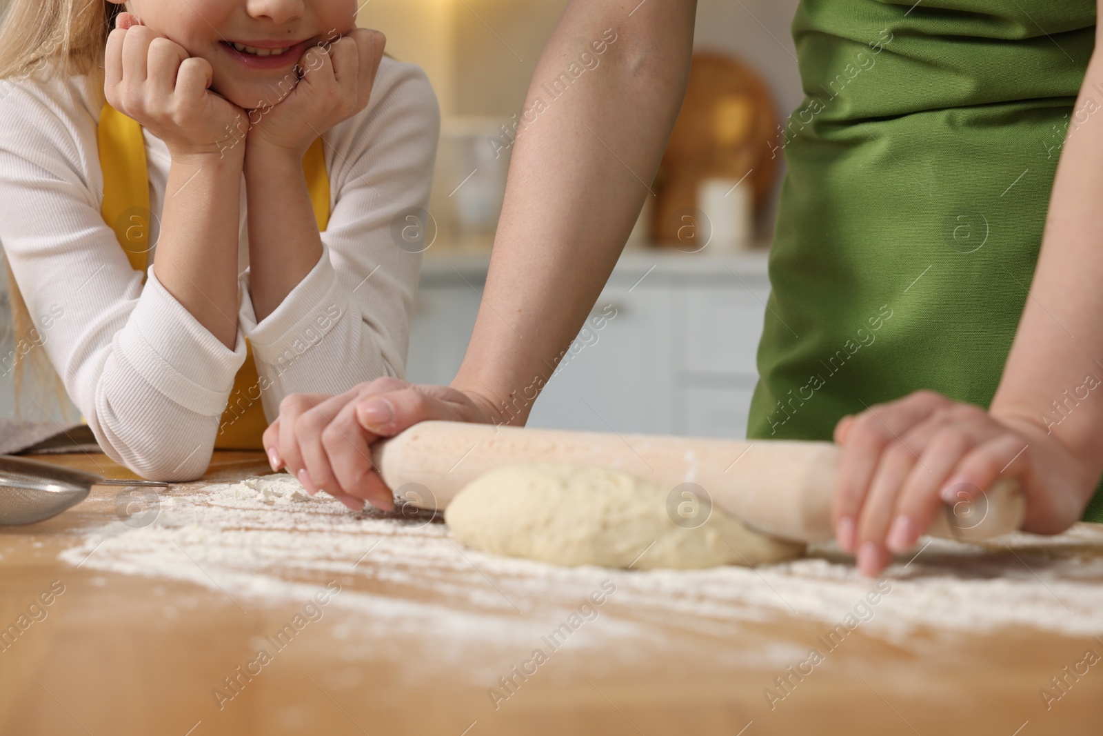 Photo of Making bread. Mother and her daughter rolling dough at wooden table in kitchen, closeup