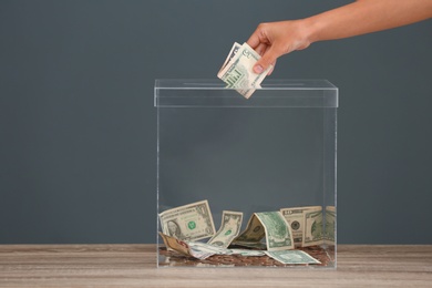 Photo of Woman putting money into donation box on table against grey background, closeup